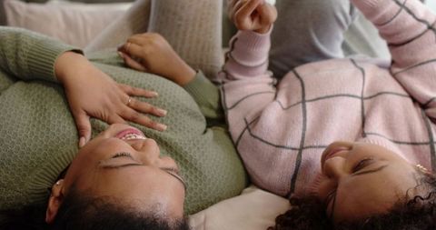 African American friends lying on bed sharing cozy laughter wearing soft knit sweaters