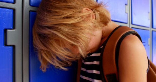Child with blond hair rests head against blue school lockers, expressing sadness or distress. Suitable for topics on childhood emotions, bullying, school-related issues, mental health, and education. The backpack and striped shirt hint at the school environment, offering relatable conditions for articles, promotions, or informational materials on children's well-being.