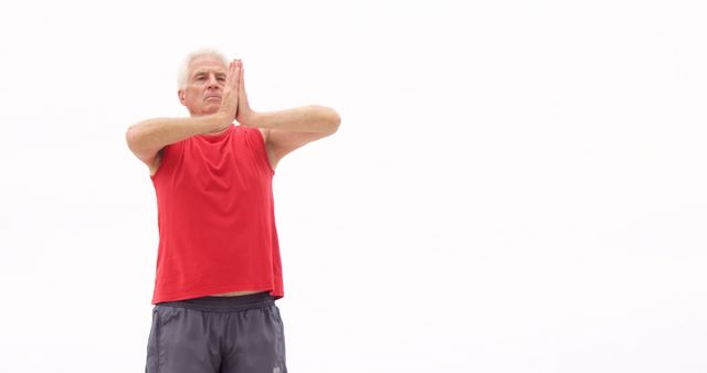 Senior man wearing red shirt and gray shorts, practicing yoga with hands in prayer pose against a white background. Ideal for concepts related to senior fitness, mindfulness, meditation, healthy lifestyle, elder care, and promoting physical health and wellbeing among older adults. Can be used in articles, social media posts, websites, and promotions focusing on senior wellness and health benefits of yoga.