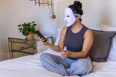 Woman wearing led facial mask on bed holding remote for at-home skincare treatment