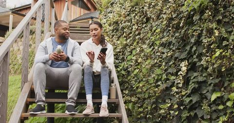 Couple sitting on wooden stairs chatting over smartphone and water bottles in backyard