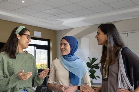 Diverse female coworkers chatting and collaborating in bright office lounge with hijab