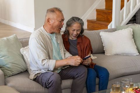Senior asian couple sharing smartphone on sofa in cozy modern living room