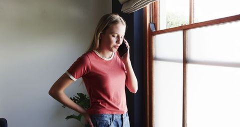 Young woman talking on smartphone by sunlit wooden window in casual home interior
