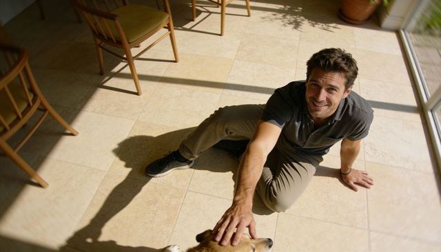 Smiling man petting dog on sunlit tile floor by glass doors in casual home interior