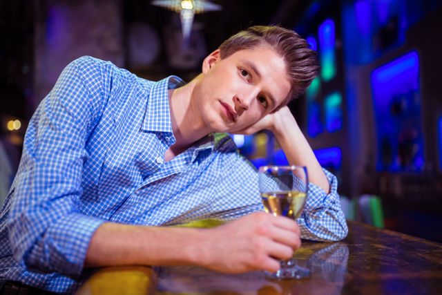 Young man sitting alone at a bar counter, holding a glass of drink, looking sad and contemplative. Ideal for use in articles or advertisements about loneliness, nightlife, mental health, or social issues. Can also be used in blogs or websites discussing the effects of alcohol or the social dynamics of bars.