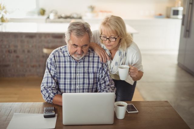 Senior couple using laptop in kitchen, both focused on screen. Man sitting, woman standing with coffee mug. Ideal for retirement planning, technology use among seniors, and lifestyle themes.