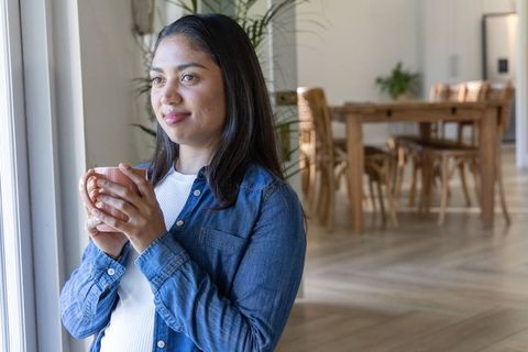Asian woman standing by window holding pink mug in cozy sunlit dining room, calm morning