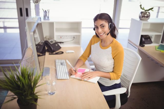 This image depicts a female customer service representative smiling while working at her desk in an office. She is wearing a headset and writing in a notebook, indicating active engagement in her tasks. This image can be used for business websites, customer service training materials, corporate presentations, and articles about workplace environments or communication.