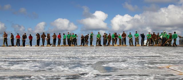 Large group of workers participating in deck cleaning task under blue sky with clouds. The lined-up team shows the importance of coordination and joint effort. Useful for illustrating concepts of teamwork, collaborative job tasks, industrial work environments, and maintenance work settings.