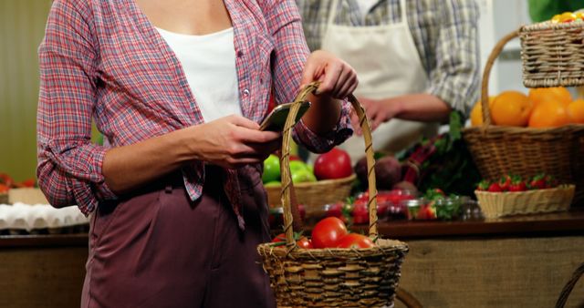 A woman is buying fresh produce at a farmers market, holding a basket filled with tomatoes. This market setting features baskets of various fruits and vegetables, with another person in the background helping and wearing an apron. Ideal for use in articles about healthy eating, organic produce, local markets, and fresh food shopping.