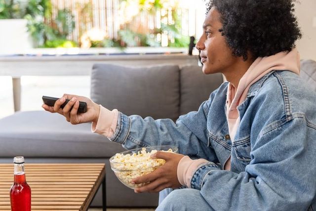 African american man watching tv at home holding popcorn and remote wearing denim jacket