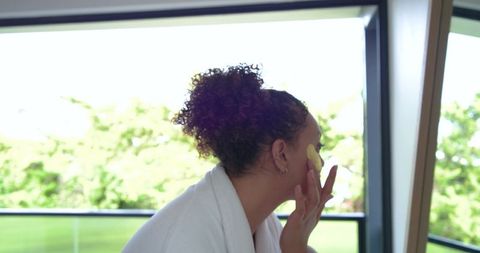 Woman applying facial pad in bathrobe by window for morning skincare and natural glow