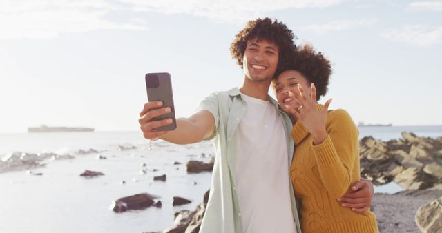 Happy couple celebrating engagement, smiling and taking selfie on rustic beach with ocean in background. Perfect for themes related to romance, togetherness, engagements, relationships, and outdoor activities.