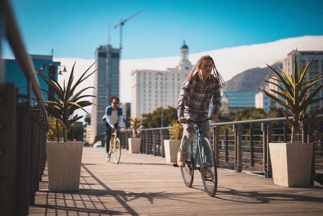 Two friends are riding bicycles on a wooden pathway in an urban cityscape. Tall buildings and a construction crane are visible in the background, along with potted plants lining the pathway. This image is ideal for promoting healthy living, urban lifestyle, outdoor activities, and friendship. It can be used in advertisements, blogs, and articles related to city life, fitness, and leisure activities.