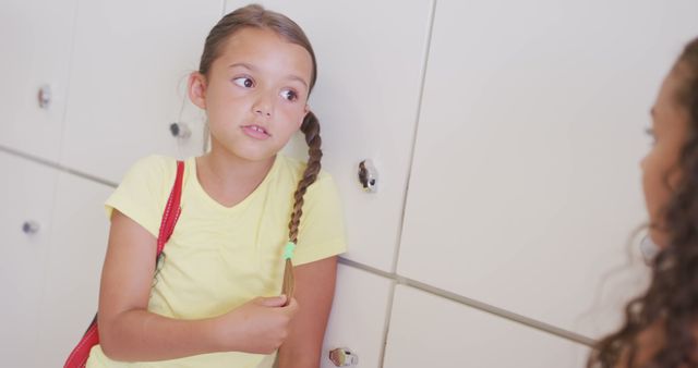 Young girl with braided hair wearing a yellow shirt, holding her hair, and talking to a friend in a school hallway near lockers. Suitable for depicting elementary school life, children's friendship, education themes, and casual student interactions.