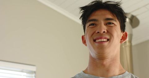 Young asian man smiling and looking up in bedroom with natural light and soft expression
