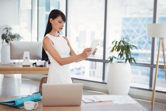 Businesswoman stands at her desk in a modern office, using a digital tablet. Ideal for illustrating business, technology, and corporate working environments, as well as showcasing modern office design and professional attire.