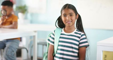 Smiling schoolgirl standing with backpack while classmate exploring with magnifying glass