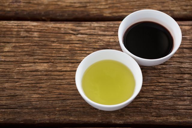 This image shows a close-up view of olive oil and balsamic vinegar in white bowls placed on a wooden table. It is ideal for use in culinary blogs, recipe websites, cooking magazines, and health food promotions. The rustic wooden background adds a natural and organic feel, making it suitable for advertisements and articles focusing on Mediterranean cuisine and healthy eating.
