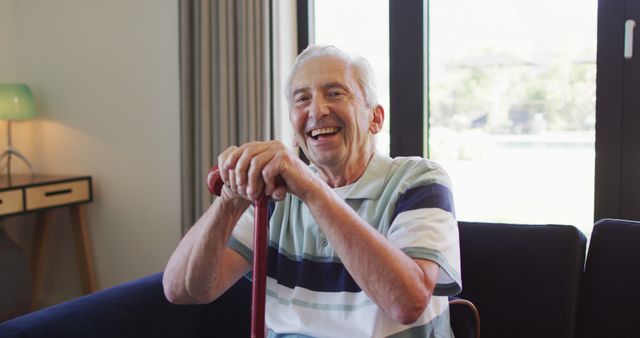 Senior man sits on couch holding walking stick and smiling in living room. Ideal for content related to elderly care, retirement living, happiness, senior lifestyle and health, family homes, and positive aging.