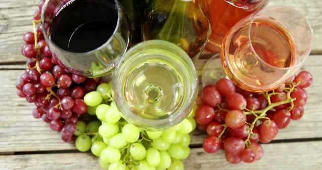 Several wine glasses filled with red, white, and rosé wine are placed along with bunches of red and green grapes on a rustic wooden table. Ideal for use in articles on wine tasting, beverage blogs, restaurant promotions, or vineyard advertisements.