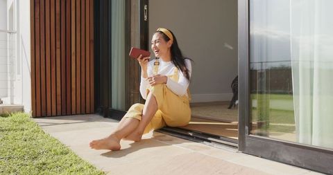 Sunlit asian woman sitting on patio laughing while holding smartphone in yellow overalls