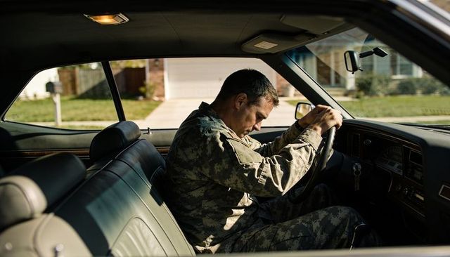 Soldier resting head on steering wheel in sunlit suburban driveway reflecting fatigue