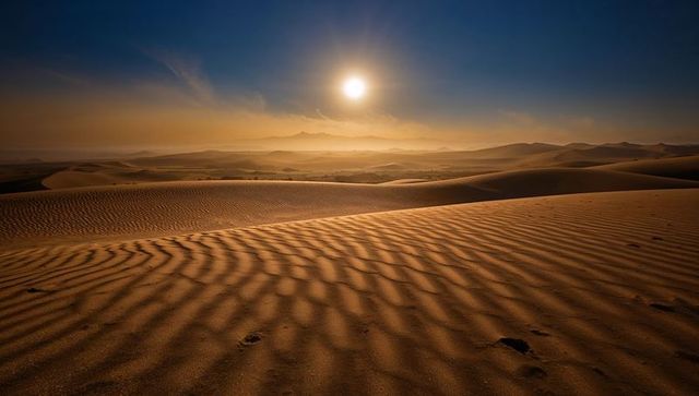 Golden sun setting over rippled sand dunes with footprints leading across desert