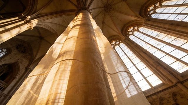 Sunlit gothic column draped in translucent fabric rising toward vaulted cathedral windows