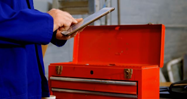 A technician in a blue uniform is using a tablet, with copy space. The open red toolbox suggests a work environment where maintenance or repairs are being performed.