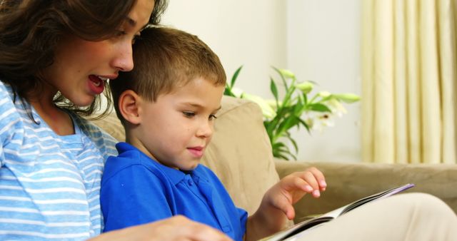 A mother is reading a storybook with her young son on a couch. They are both engrossed, with the child focusing on the book. This image conveys themes of family bonding, education, and nurturing. It can be used in advertisements for educational products, family services, or parenting articles to showcase family engagement and learning activities.