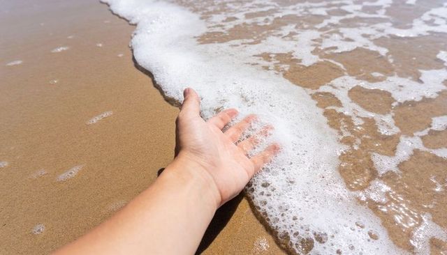 Reaching hand touching sunlit shoreline foam on sandy beach, palm feeling wave
