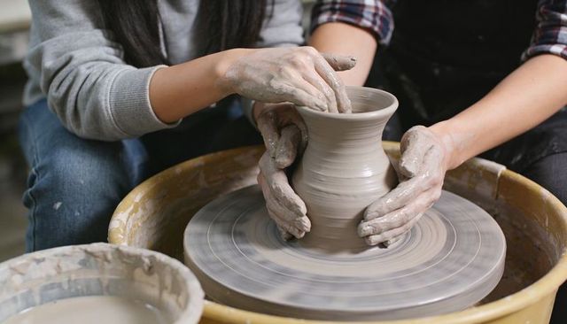 Instructor guiding student shaping clay vase on pottery wheel, hands covered in wet clay