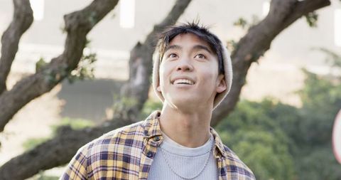 Smiling asian man looking up in urban park wearing beanie and plaid flannel shirt