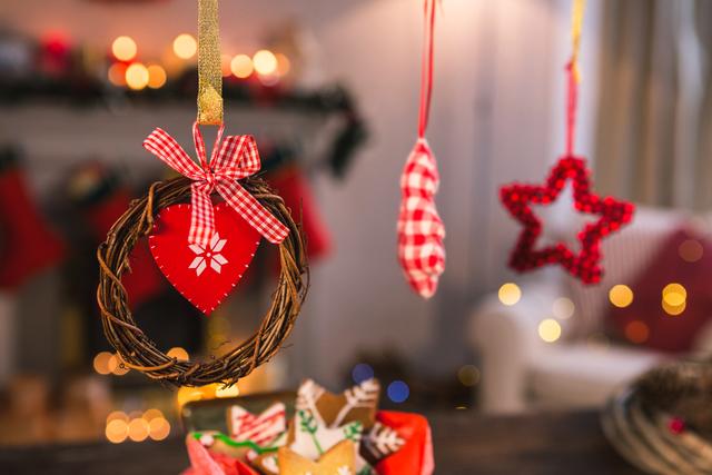 Christmas ornaments and gingerbread cookies create a festive atmosphere in a cozy living room. The image features a heart-shaped ornament with a red plaid ribbon, a star-shaped ornament, and a small wreath. The background includes warm lights and holiday decorations, perfect for use in holiday marketing, greeting cards, or seasonal blog posts.