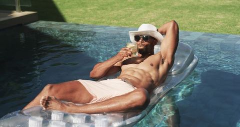 Reclining man relaxing on clear pool lounger wearing white hat and sunglasses, sipping drink
