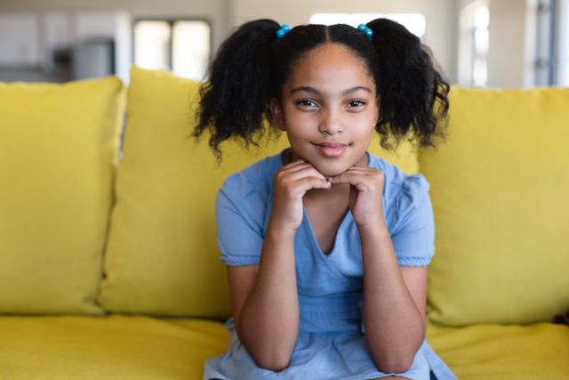Young biracial girl in blue dress sitting on yellow sofa in school playroom, smiling with hands on chin. Ideal for educational materials, childhood development articles, school brochures, and advertisements promoting a positive school environment.