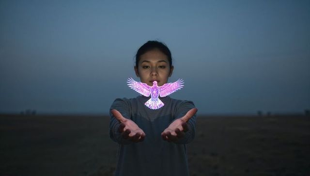 Asian woman holding glowing holographic bird at twilight in open field, neon magic