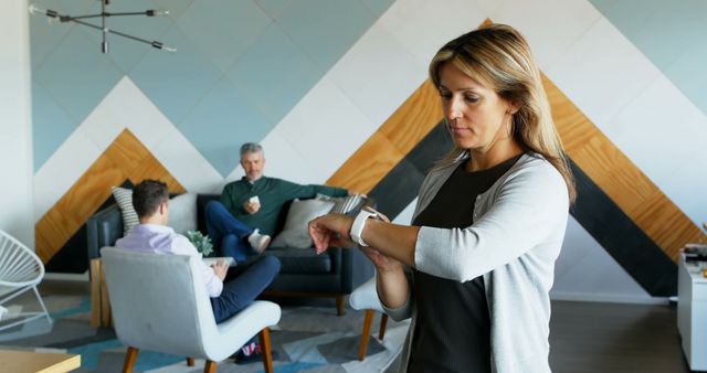 Businesswoman checking smartwatch in modern office while two male colleagues converse in the background. Ideal for business, technology, time management concepts, showcasing modern workplace environments, and team collaboration.