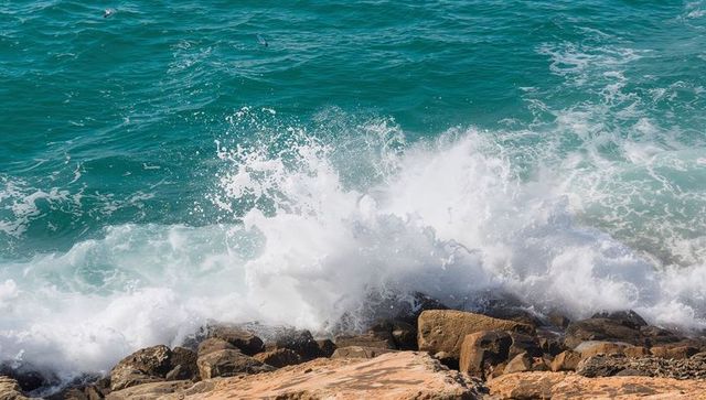 Turquoise wave crashing over sunlit coastal rocks with dramatic sea spray and foam