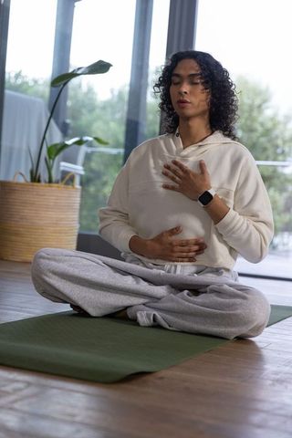 Non-binary person meditating on yoga mat by glass doors wearing smartwatch for mindfulness