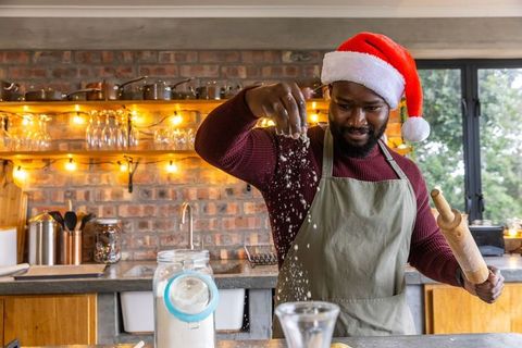 African american man wearing santa hat sprinkling flour while baking in cozy kitchen