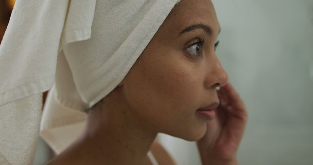 Closeup profile of a woman with a towel wrapped around her head, suggesting she has just taken a shower. She is looking away, touching her face, capturing a moment of calm and self-care. Ideal for use in wellness, beauty, skincare advertisements or articles focusing on personal care and spa treatments.