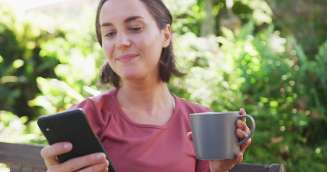 Smiling caucasian woman using smartphone and holding mug of tea sitting in sunny garden. domestic life and leisure time concept.