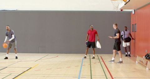 Five men practicing basketball drills on indoor hardwood court with hoop, towel, duffel