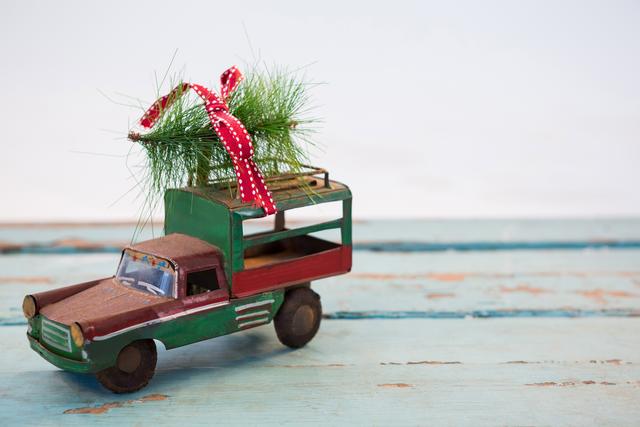 Vintage toy truck carrying a small Christmas tree tied with a red ribbon, placed on a weathered wooden plank. Ideal for holiday-themed decorations, greeting cards, festive advertisements, and nostalgic Christmas promotions.
