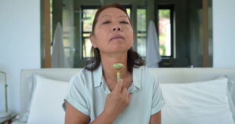 Senior asian woman using jade roller on neck while sitting on bed for morning self-care