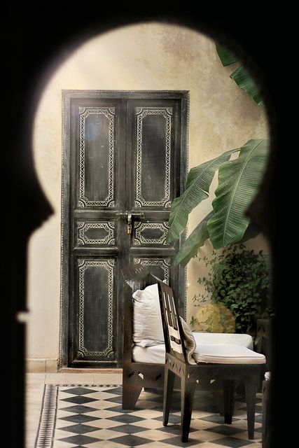 Moroccan-style doorway with intricate designs viewed through a keyhole. Includes a wooden chair with a cushion, tropical plants, and a decorative checkerboard floor. Ideal for use in travel magazines, cultural blogs, interior design inspiration, and architectural features.