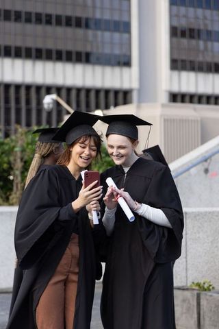 Graduates celebrating on urban steps sharing smartphone and diplomas, friends smiling